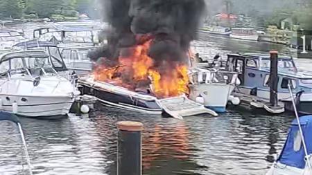 A blurry screenshot from CCTV footage showing a dark blue boat moored up to a pontoon, with a fire raging on most of the deck and cockpit with black smoke billowing into the air