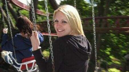 Gemma Sisley is sitting on a swing, her head is turned towards the camera and she is smiling. She is wearing a black hoody.