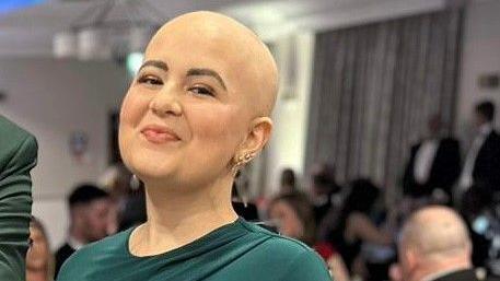 A woman who has lost her hair from cancer treatment smiles at the camera. She's wearing a green dress and has several gold earrings in her left ear. Behind her is a crowd that appears to be at some sort of dinner event.