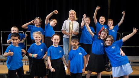 A group of 12 primary school children wearing blue t-shirts, stood on a stage with a woman in a white top holding a gold Olympic torch from the 2012 London Olympics. 