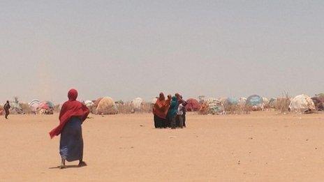 A woman walks across a sandy stretch of land towards a group of people, with a camp of tents behind them.