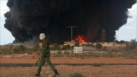 A rebel walking past a burning oil terminal