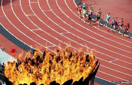 Athletes in steeplechase heats run pas the Olympics cauldron