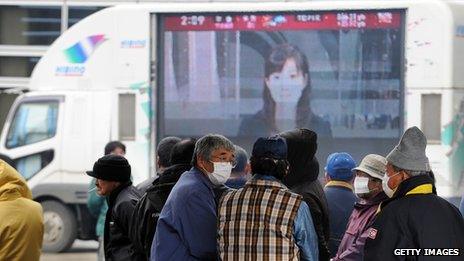 Japanese people stand in front of a large TV screen showing Japanese news