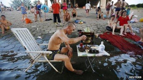 A man sitting on a beach chair in his swimming trunks takes his meal at a small table partially dipped into the Hanjiang river to escape the summer heat, as other swimmers look on under a bridge in Wuhan, Hubei province, 28 July 2013
