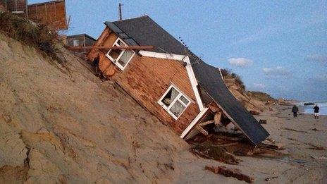 A wooden building which has slipped down a sand bank onto a beach