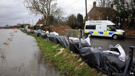 A police van parked behind temporary flood defences at a river bank