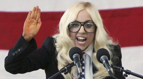Lady Gaga dressed in blonde wig and wearing suit speaking at a rally