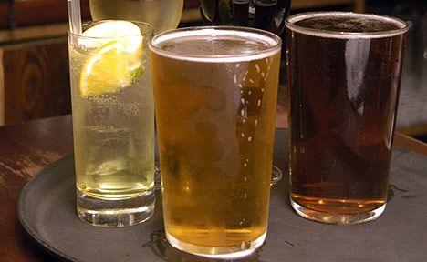 Three drinks on a tray on a pub table.  From left to right they appear to be a spirit and mixer with ice, a slice of lemon and a straw, a pint of lager and a pint of ale.