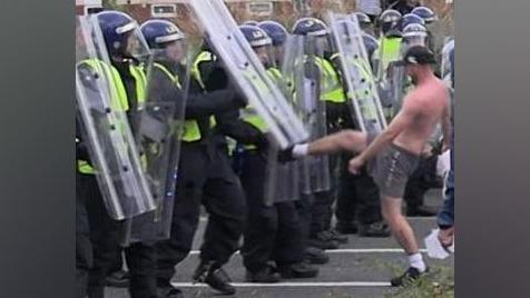 A shirtless man, wearing a black cap and shorts, kicks under the shield of one of a line of police officers in riot gear.