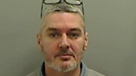 Mugshot of a man with short grey hair and a trimmed bear and glasses on his head, he is wearing a grey sweatshirt