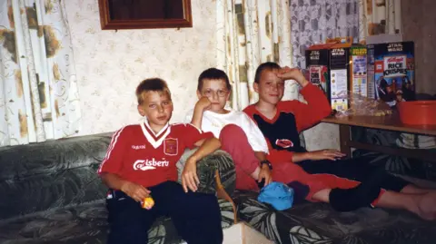 Anton Honchanka Dimtry on the left wearing an old Liverpool football top. Two boys in pyjamas sit on the right of him. There is a table on the right with cereal boxes.