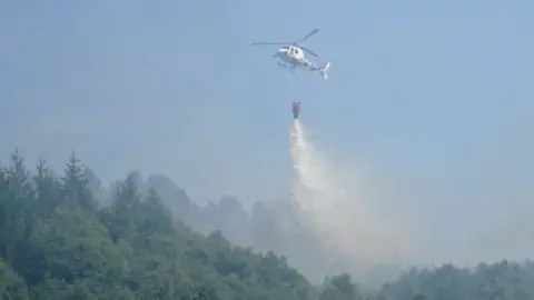 @SWPCynon/Twitter A helicopter dropping water on the blaze near Bryn Pica landfill site in Aberdare