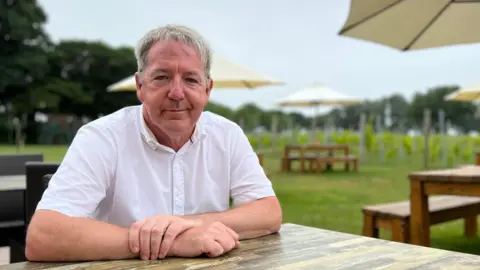 Tim sits at an outdoor wooden table, wearing a white shirt with both arms resting on the surface. The background features large umbrellas shading other tables and benches, set in a grassy area with trees and what appears to be a vineyard in the distance.