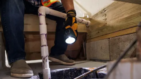 A man in dark blue jeans, high vis and gloves shining a torch down the hole where a pipe is, looking for damage.