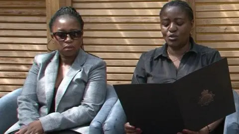 GMP Two female relatives of Julius Matera, both with black braided hair and one wearing a grey jacket and sunglasses and the other a black shirt and holding a large black folder with a police crest on it, sit on blue chairs in front of horizontal-slated wooden fencing.