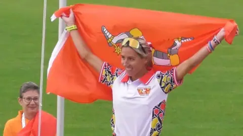 Christa Cain is on top of a podium. She is wearing the Isle of Man team top of a white polo shirt with multi-coloured swirls on the shoulders, along with black trousers and gold-coloured sunglasses on her head. She is holding up a red flag Manx flag behind her, and smiles. A woman in an orange t-shirt looks on.