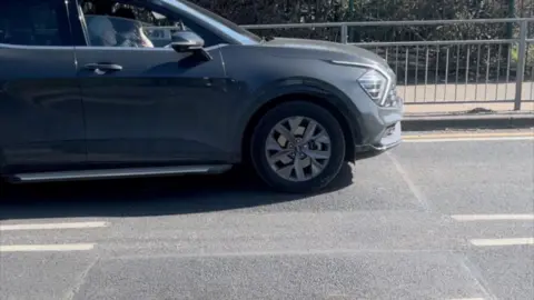 Elliot Deady/BBC A grey car driving over a speed bump. It is driving from left to right. There are metal railings and a bush in the background.