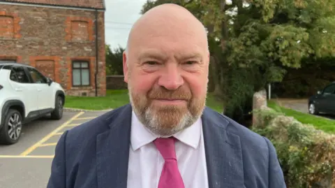 A man in a navy suit and pink tie looking at the camera with a neutral expression and standing in a car park