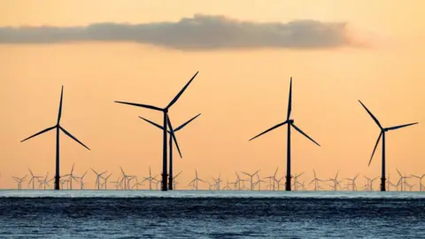 A large offshore wind farm with a number of turbines quite close, with tens of them in the distance, against an orange sky.