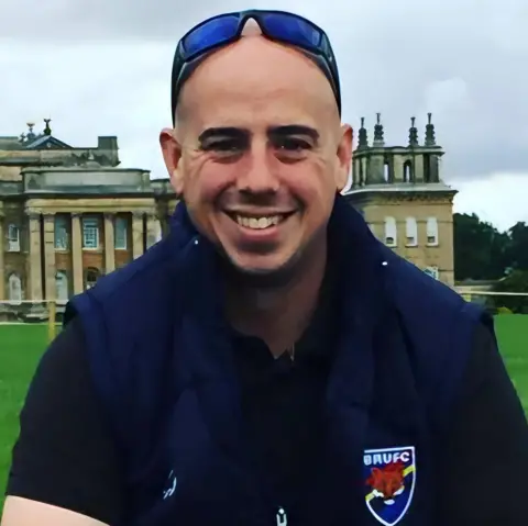 Charlie Sadler Martyn Sadler with stately home behind him - he is wearing a dark blue rugby top and has sunglasses on his head. He is looking directly at the camera and smiling.