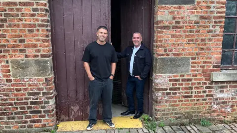 Two men stand in the doorway of an old millhouse. One is wearing a black t-shirt and jeans, while the other is wearing a grey jacket atop a white t-shirt. Both are smiling into the camera.