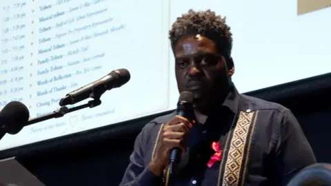 Sam Read/ BBC Jabu Ncube stands with a microphone in one hand on a stage. 