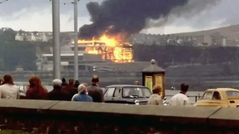 NOEL HOWARTH A group of people look on as the fire takes hold of the Summerland complex with black smokie rising above it.