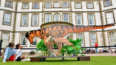 A large striped black and orange dinosaur model made from Lego bricks, a woman and a boy are sitting on the lawn outside Sewerby Hall stately home looking up at it.