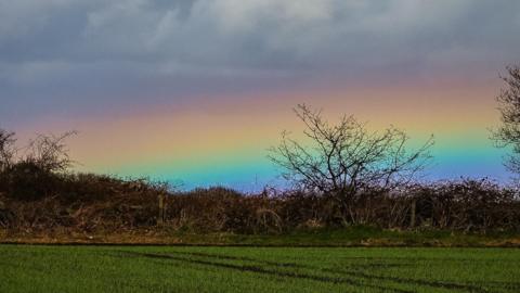 Cloudy sky with the edge of a rainbow above green fields.