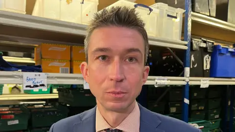 Peter Edwards, a man with light brown hair dressed in a blue suit and tie with a red striped shirt, looks into the camera in a warehouse.