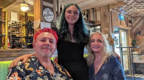 Supplied Davel Llewellyn with his daughter Amy and wife Ange. They are sitting in a pub and smiling at the camera. Dave has red spiked hair, Amy has long green hair and glasses and Ange is wearing a sparkly top and has blonde hair.