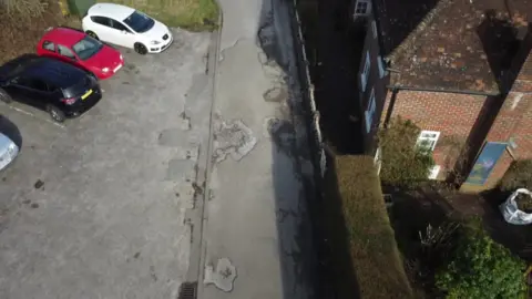 A bird's eye view of a road covered in potholes. To the left are four parked cars next to a grass verge and some hedging. To the right of the road is a metal fence and red brick houses with box hedging and a garden just in view. The potholes take up half of the section of road.