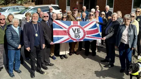 Crowthorne Parish Council A group of about 30 people gathered outside a village hall. The people at the front are holding up a VE Day 80 flag which is a union flag with a circle of poppies in the middle. Inside the poppies are the words, VE Day 80.