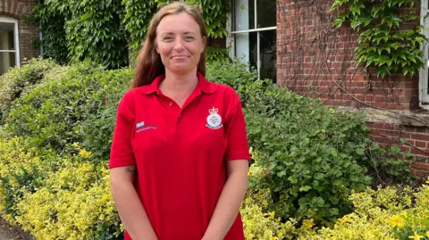 Clinical nurse specialist Vicki Bailey stands in front of some bright green shrubs. She is wearing a red polo shirt and has long ginger hair. 