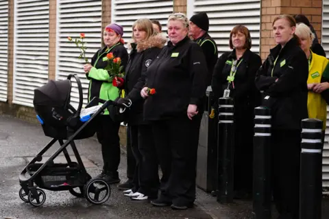 PA Media A number of workers in Asda uniforms stand on a pavement. Some are tearful, others are holding orange roses. One has a pram.