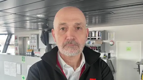 Duncan Mackison is a bald man with grey facial hair. He wears a white shirt and black fleece and stands in the bridge of a ferry