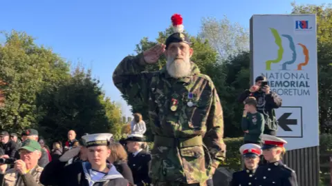 A man in camouflage, wearing a beret with a red and white plume, salutes, with others in military uniform stood around him 