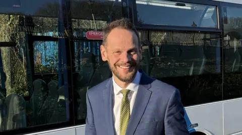 Simon Lightwood wearing a navy blue suit and gold tie and white shirt standing smiling with his hands clasped together in front of a white bus.