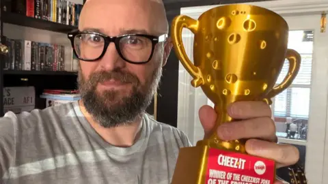 A close-up indoor photo of Fil Milton wearing large black-framed glasses and a grey T-shirt, holding up a shiny gold trophy towards the camera. He has a brown beard. The trophy has a red label reading "Cheez-It" and includes the words "Winner of the Cheeziest Joke." Bookshelves filled with books and DVDs are visible in the background.