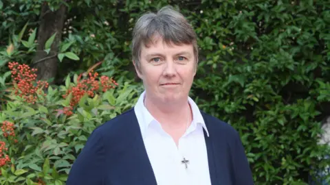 A woman is wearing a cross around her neck. She's standing outside with bushes behind her. She has a white short and a navy cardigan on. She has short hair. 
