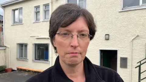 A lady with short dark hair stands in front of a white building. Sh has a sombre expression, is wearing eye galssses, a black coat and grey and white top