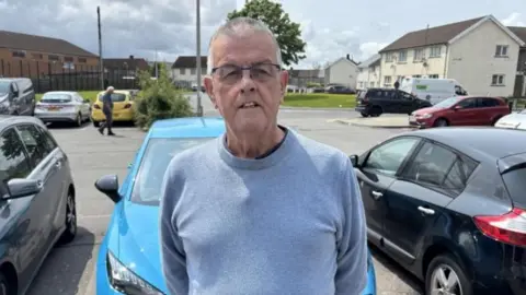 Steve Collins standing in front of a line of parked cars wearing a light blue shirt. Housing, grass verges and a tree covered in green leaves are in the background, with the tree partly obscured by Steve's head.