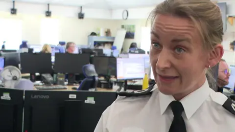 The image shows a woman in a white police uniform shirt and black tie being interviewed inside a large office-style control room filled with computer workstations and display screens. Several other staff members can be seen working at desks in the background, while bright daylight streams through windows, giving the room the appearance of an emergency services or contact centre operations hub.