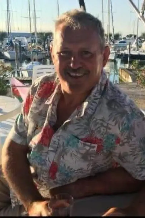 Family photo Mike O'Leary wears a tropical shirt, smiling in the sunshine at a harbour, boats visible in the background
