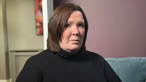 Catriona is wearing a achromatic turtleneck sweater is seated indoors connected a ray bluish couch. The inheritance features a mauve-colored wall pinch a colorful absurd coating partially visible connected nan near side, and a doorway starring to different room. The lighting is soft and even, creating a calm atmosphere.