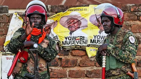Two policemen in red crash helmet stand in front of brick wall with old posters of Uganda's President Yoweri Museveni against a yellow background.