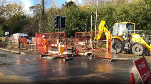 A JCB digger is pictured behind a metal fence as workmen in orange coats, orange trousers and hard hats work to fix the leak.