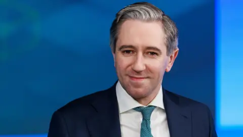 Getty Images Simon Harris is smiling. He's wearing a navy suit with a white shirt, and a blue tie decorated with white spots. His hair is black and white and brushed to the side. The background is blue and out of focus. 
