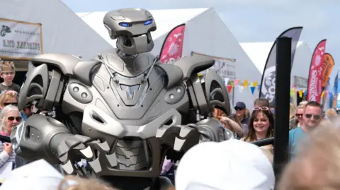 Matt Keeble/PA Wire People look at a robot that has a human-like form. It stands above their heads and is silver with blue lights for eyes. It is appearing at the Royal Cornwall Show and large white tents can be seen behind it. 